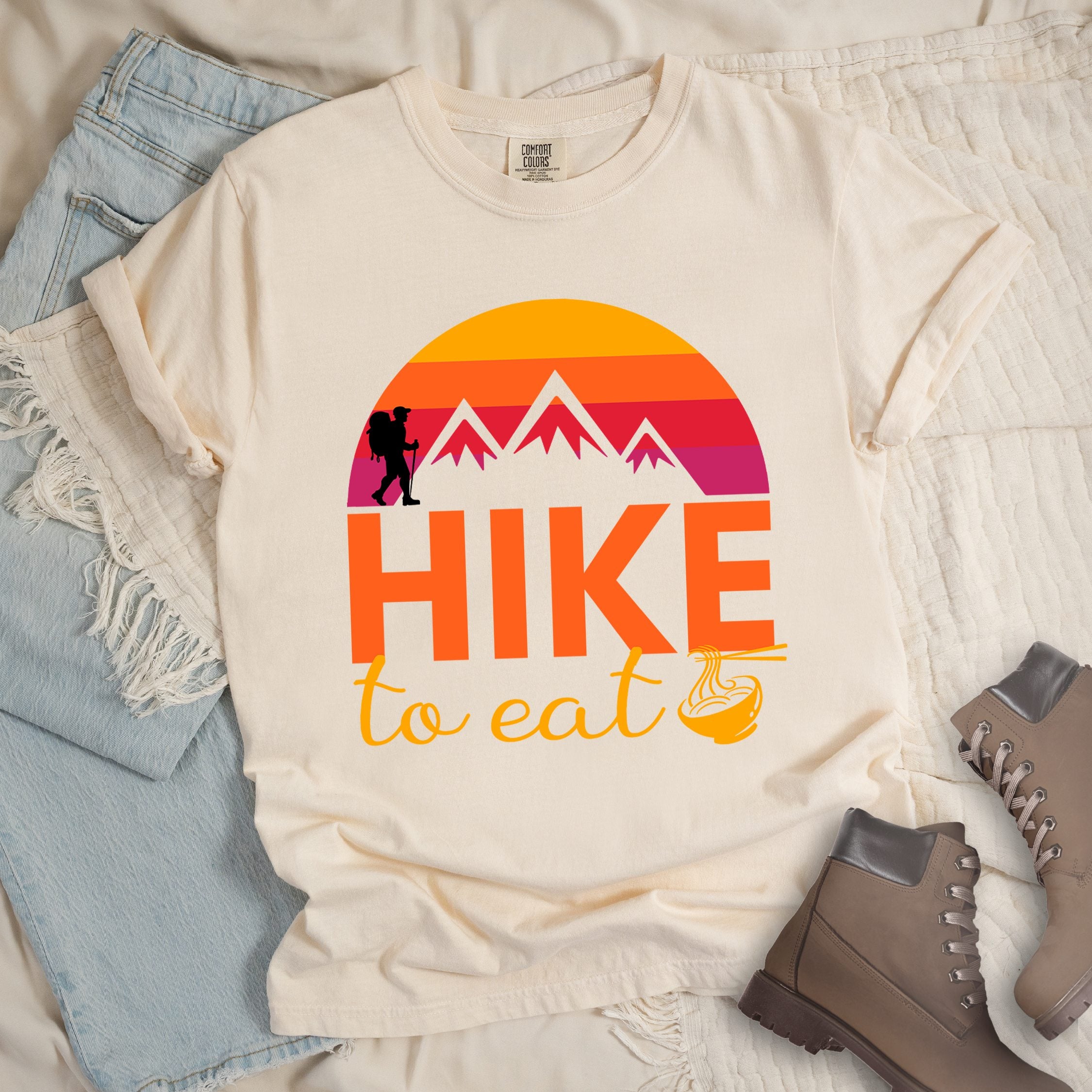 Ivory shirt with the text “Hike to Eat” and a circular mountain and sunset graphic, with a small bowl of noodles integrated into the design.