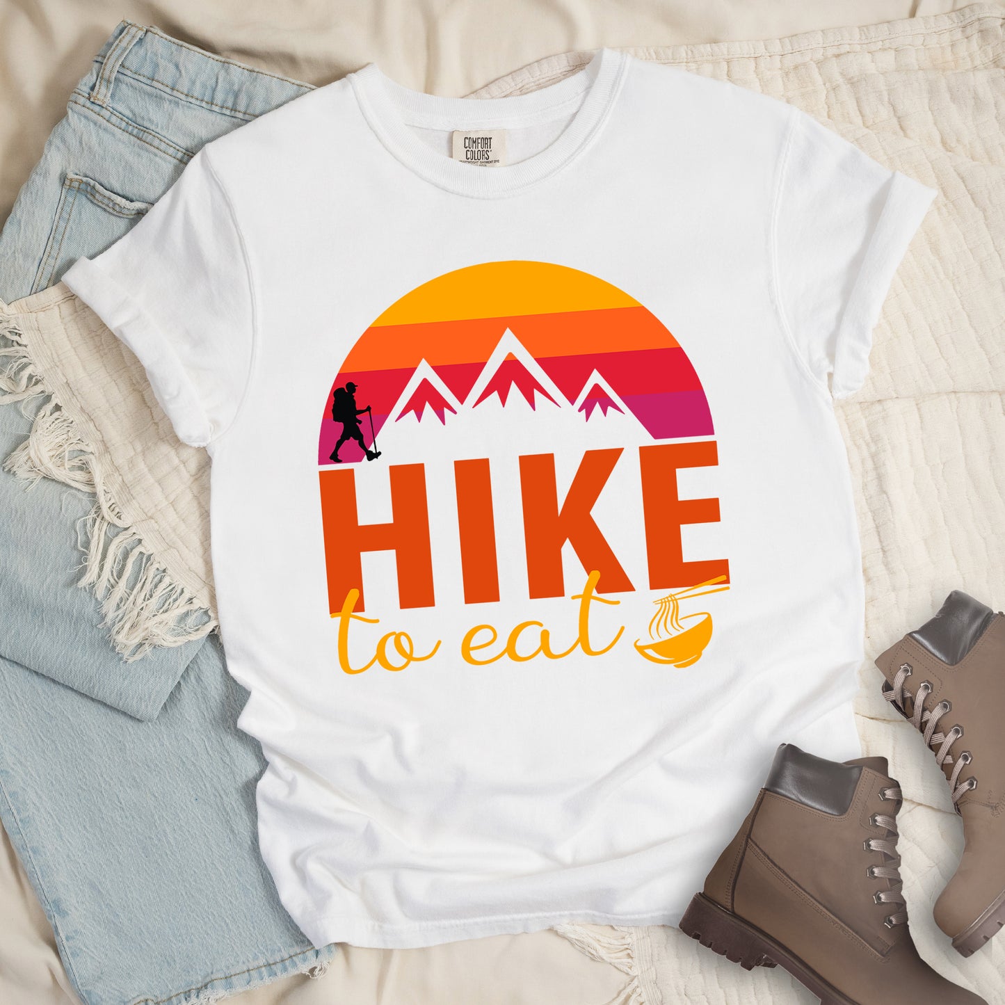 White t-shirt featuring a retro sunset design with gradient colors from yellow to orange to red to purple. Mountain silhouettes and a small hiker figure appear against the sunset. Below this is "HIKE" in large orange letters and "to eat" in yellow script, followed by a small illustration of a noodle bowl with chopsticks. The design has a vintage outdoor aesthetic. The shirt features a classic crew neck and casual rolled sleeves, styled alongside light wash denim and brown hiking boots.