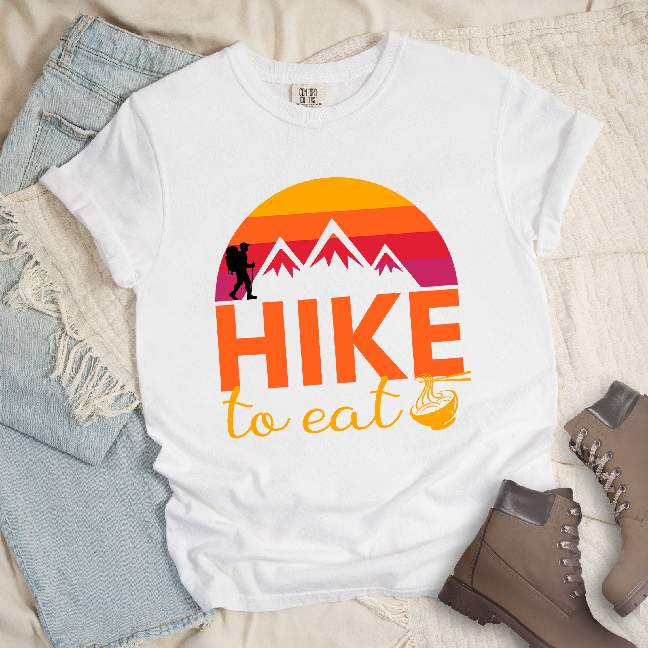 White shirt with the text “Hike to Eat” and a circular mountain and sunset graphic, with a small bowl of noodles integrated into the design.