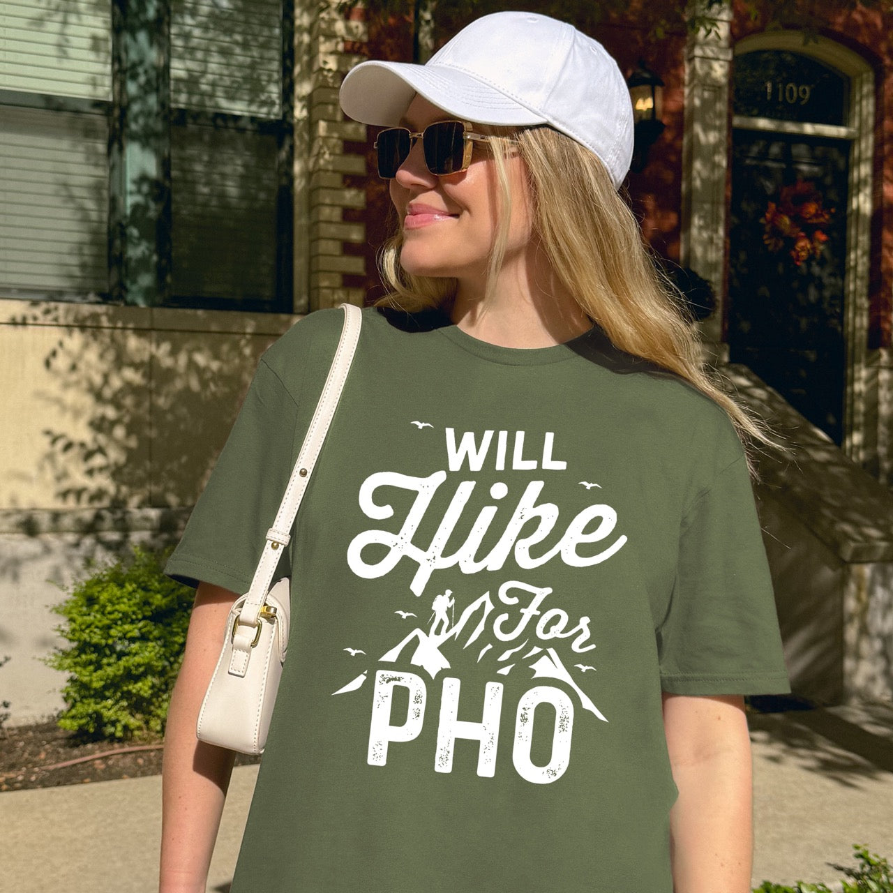 woman wearing military green Will Hike for Pho statement tee with bold and script typography, mountain peaks, hiker silhouette, and flying birds for Vietnamese noodle-loving outdoor enthusiasts, hikers and foodies 