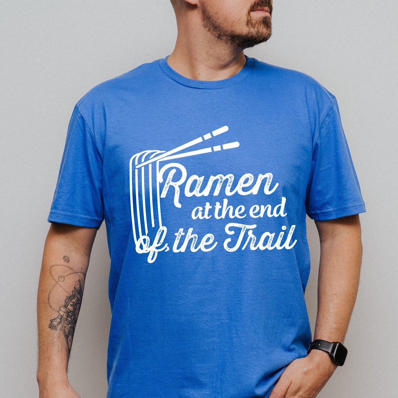 Man wearing Royal blue Ramen at the End of the Trail statement tee with elegant script typography, ramen bowl illustration, and chopsticks for Japanese noodle-loving outdoor enthusiasts, foodies and hikers