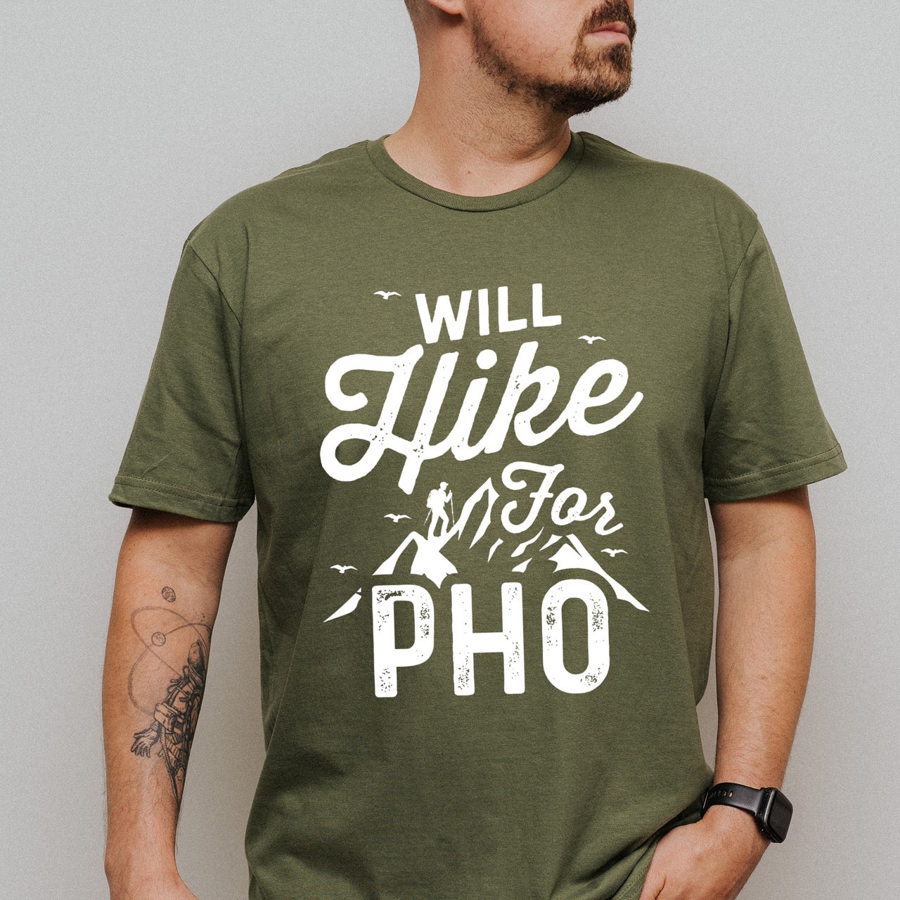 man wearing military green Will Hike for Pho statement tee with bold and script typography, mountain peaks, hiker silhouette, and flying birds for Vietnamese noodle-loving outdoor enthusiasts, hikers and foodies