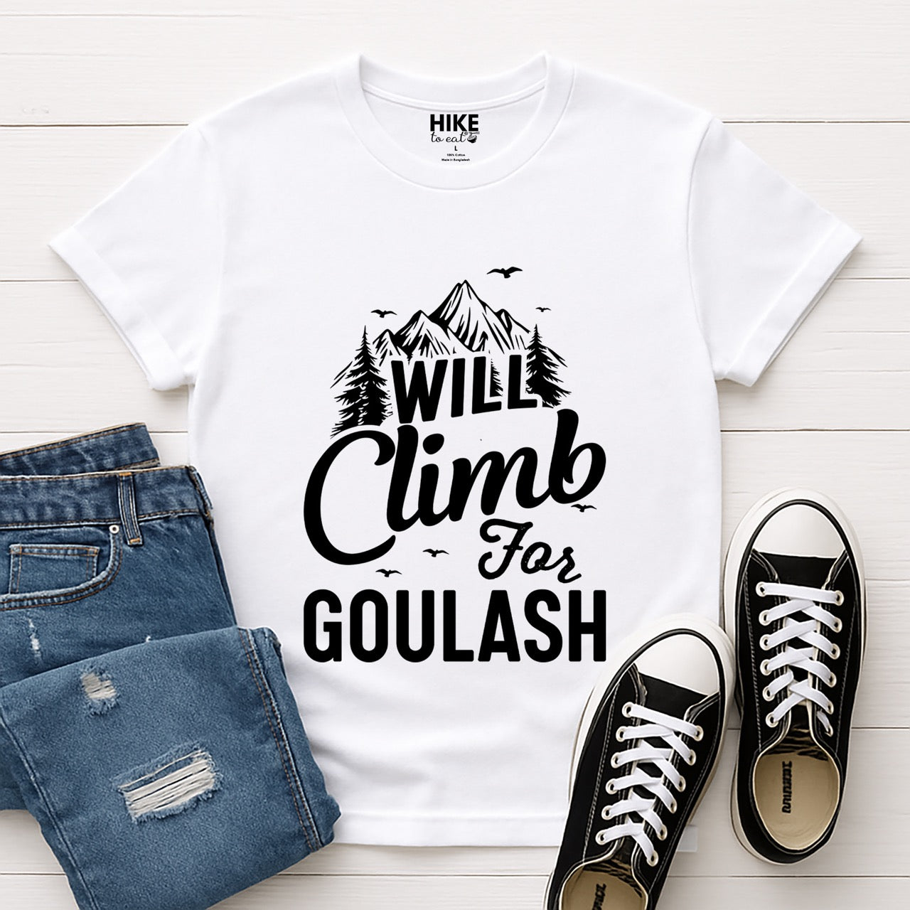 white Will Climb for Goulash statement tee with bold and script typography, mountain peaks, pine trees, and flying birds for Hungarian food-loving climbers, hikers and foodies laid flat with shoes and jeans