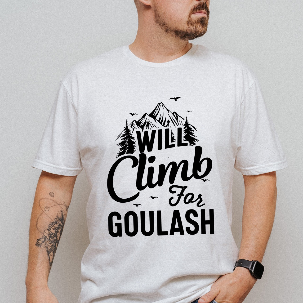 man wearing white Will Climb for Goulash statement tee with bold and script typography, mountain peaks, pine trees, and flying birds for Hungarian food-loving climbers, hikers and foodies