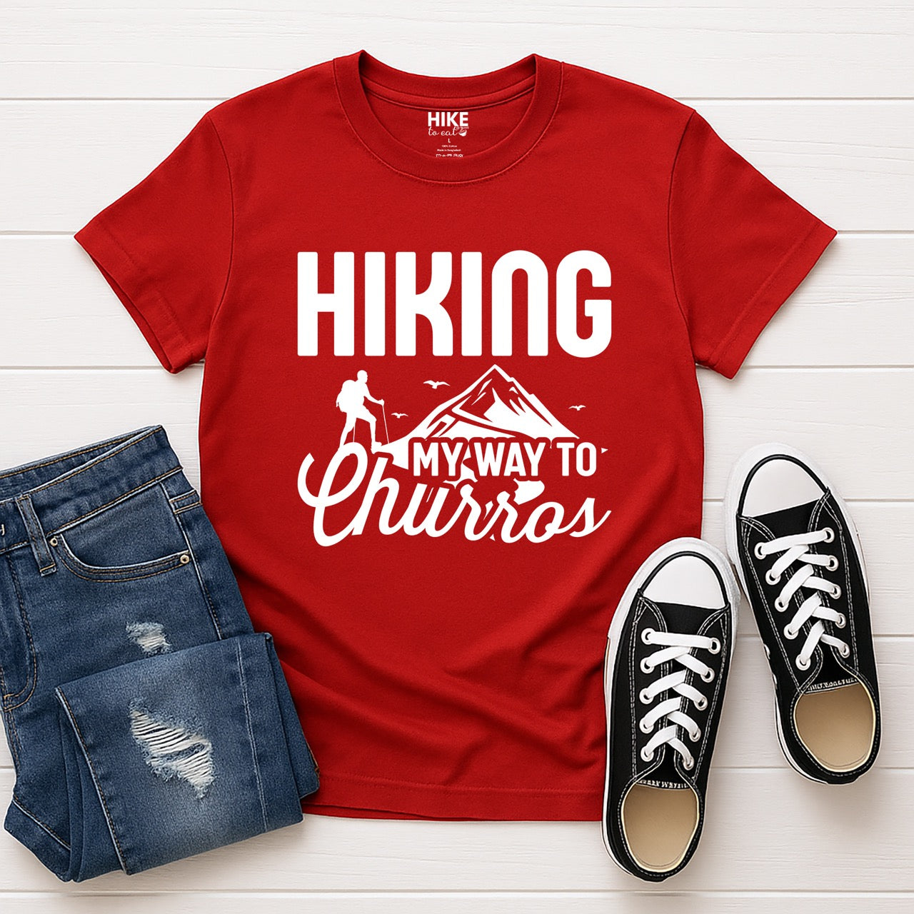 Red Hiking my way to Churros statement tee with bold typography, script lettering, mountain peaks, hiker silhouette, and flying birds for Spanish dessert-loving outdoor enthusiasts, hikers and foodies laid flat with jeans and shoes