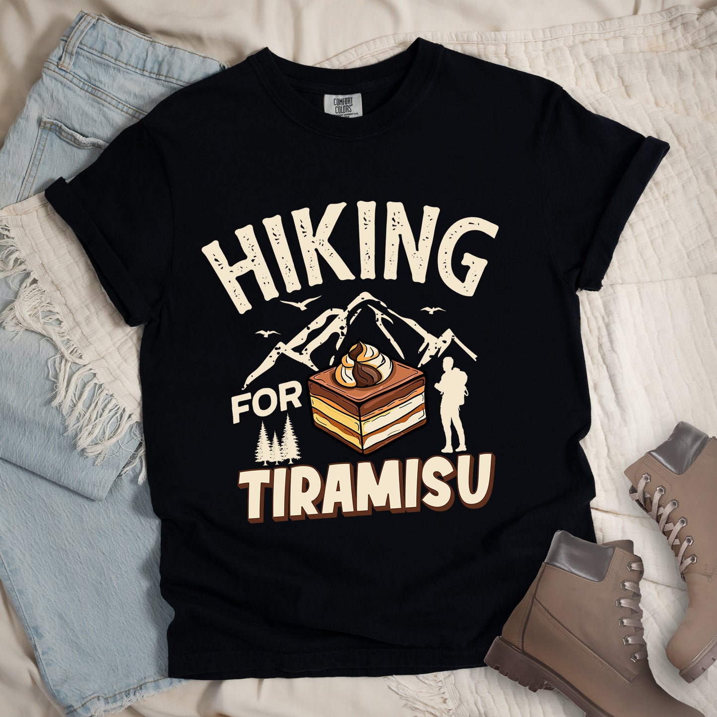 Black t-shirt with cream-colored distressed text reading "HIKING FOR TIRAMISU". The word "HIKING" appears at the top, with mountain silhouettes, small birds, and pine trees below it. A hiker silhouette stands to the right of the design. In the center is a detailed illustration of a tiramisu dessert square showing layers of coffee-soaked cake and cream, topped with a swirl of mascarpone. The word "TIRAMISU" appears in brown gradient letters at the bottom.