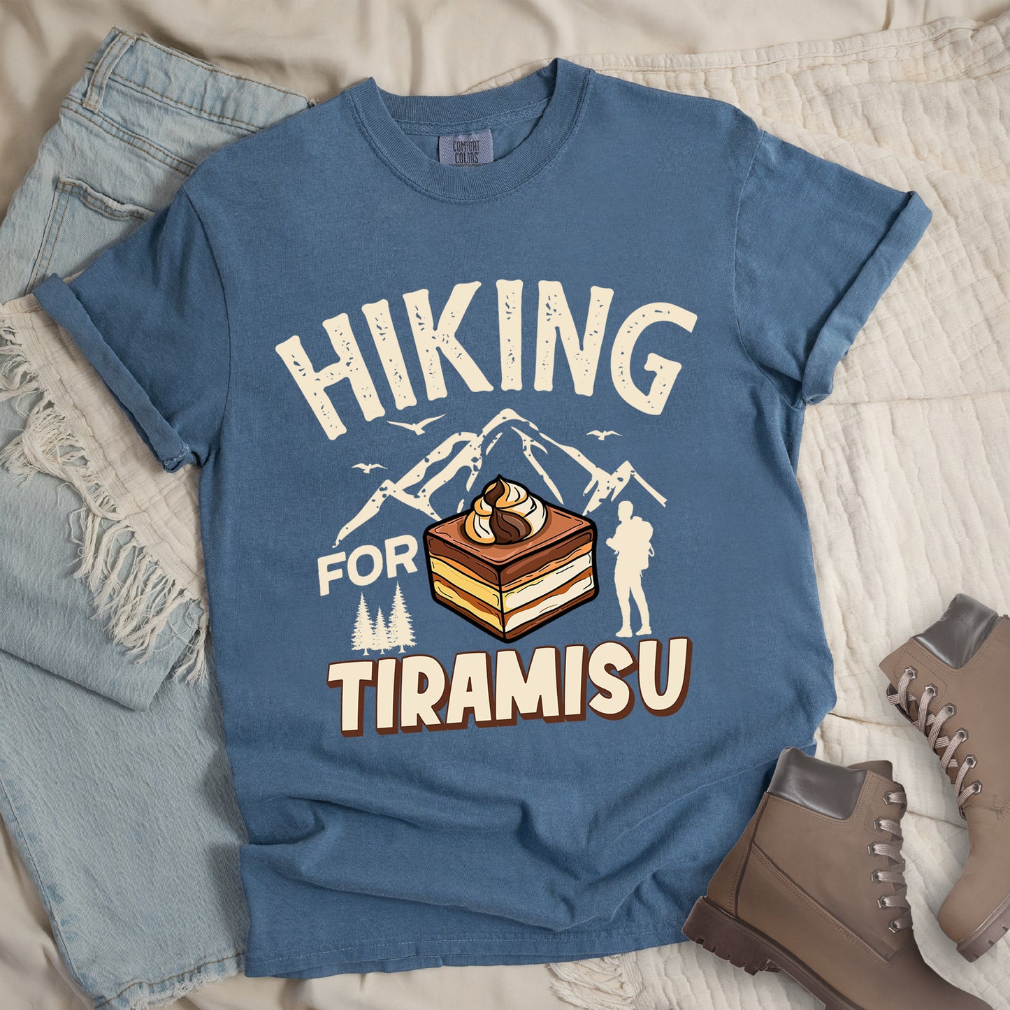 Blue Jean (Denim Blue) t-shirt with cream-colored distressed text reading "HIKING FOR TIRAMISU". The word "HIKING" appears at the top, with mountain silhouettes, small birds, and pine trees below it. A hiker silhouette stands to the right of the design. In the center is a detailed illustration of a tiramisu dessert square showing layers of coffee-soaked cake and cream, topped with a swirl of mascarpone. The word "TIRAMISU" appears in brown gradient letters at the bottom.
