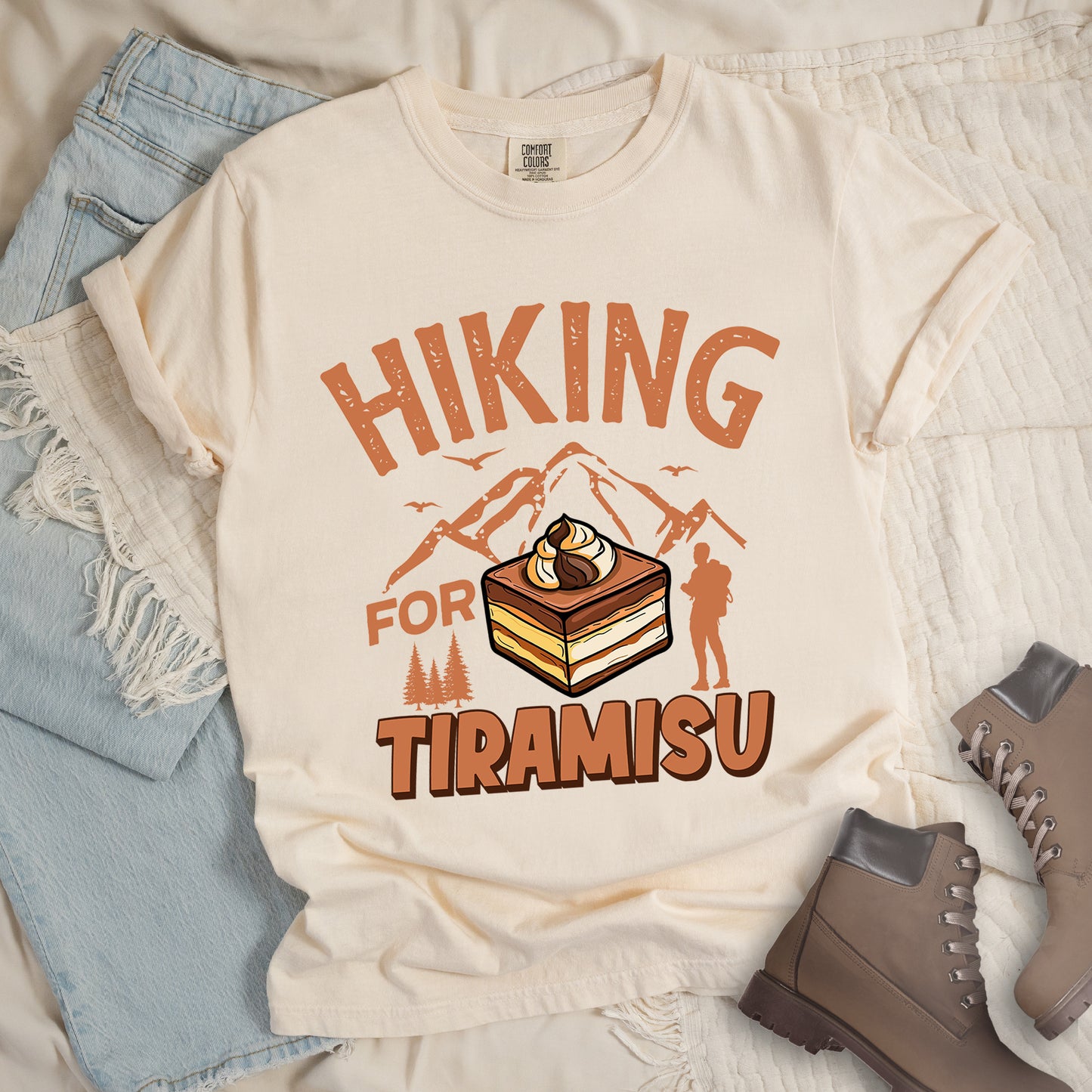 Ivory (Cream White) t-shirt with light brown-colored distressed text reading "HIKING FOR TIRAMISU". The word "HIKING" appears at the top, with mountain silhouettes, small birds, and pine trees below it. A hiker silhouette stands to the right of the design. In the center is a detailed illustration of a tiramisu dessert square showing layers of coffee-soaked cake and cream, topped with a swirl of mascarpone. The word "TIRAMISU" appears in brown gradient letters at the bottom.