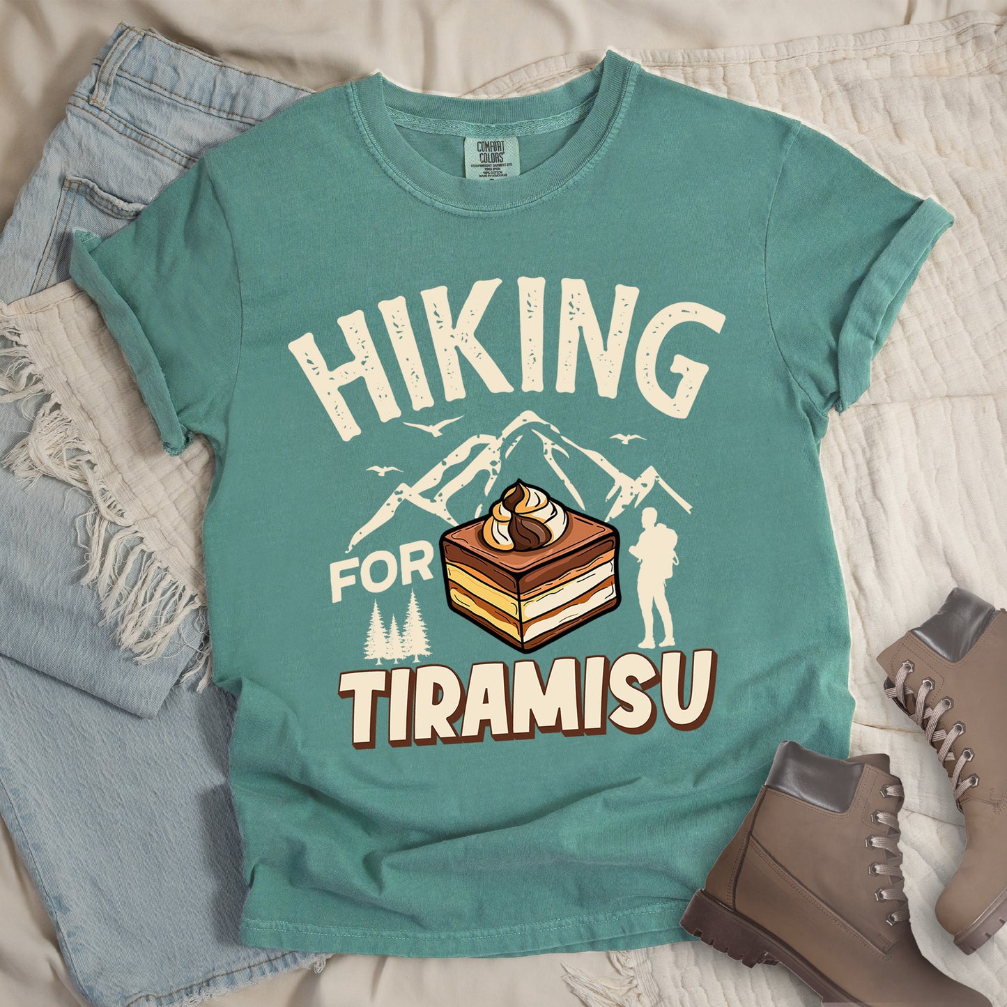 Light Green t-shirt with cream-colored distressed text reading "HIKING FOR TIRAMISU". The word "HIKING" appears at the top, with mountain silhouettes, small birds, and pine trees below it. A hiker silhouette stands to the right of the design. In the center is a detailed illustration of a tiramisu dessert square showing layers of coffee-soaked cake and cream, topped with a swirl of mascarpone. The word "TIRAMISU" appears in brown gradient letters at the bottom.