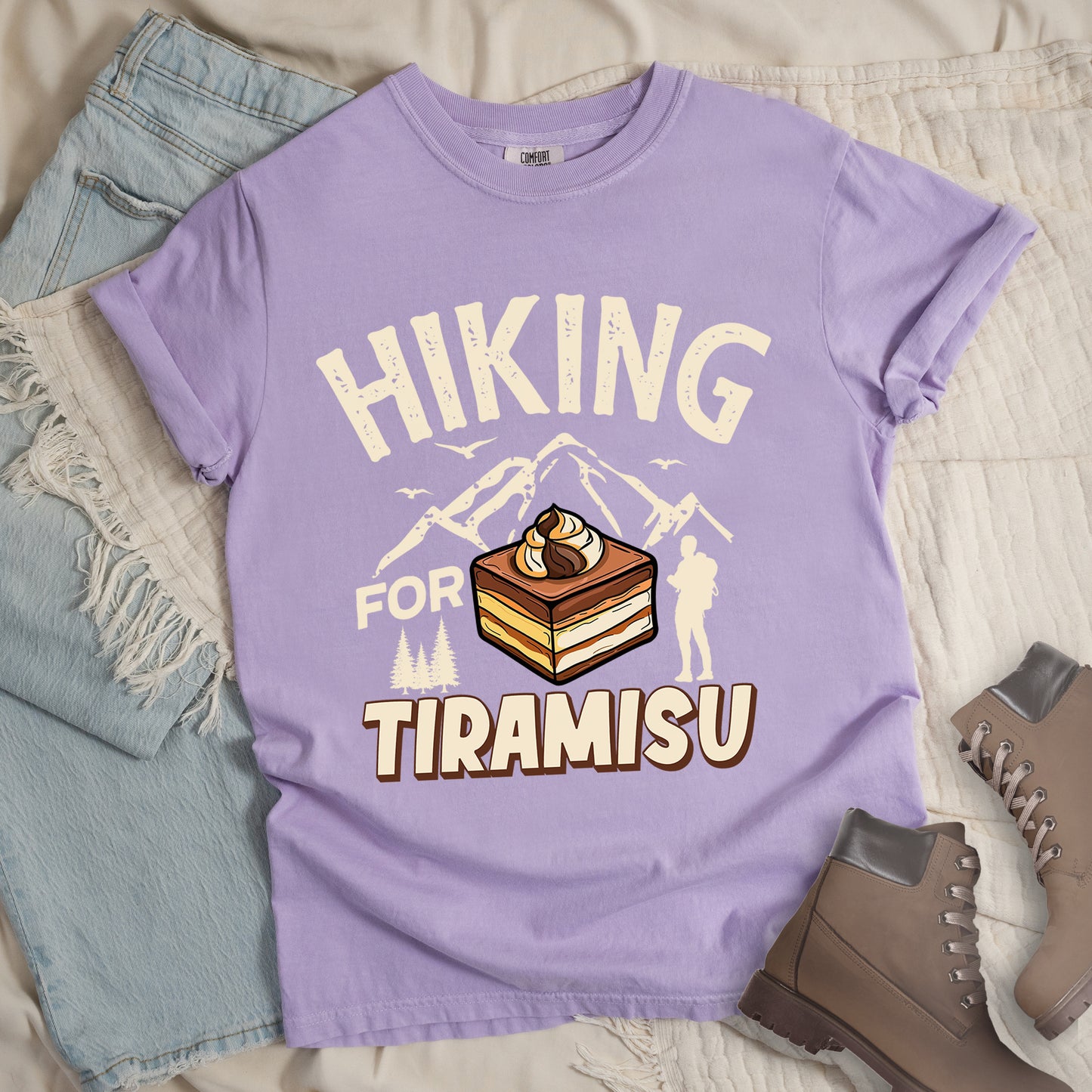 Orchid (Light Violet) t-shirt with cream-colored distressed text reading "HIKING FOR TIRAMISU". The word "HIKING" appears at the top, with mountain silhouettes, small birds, and pine trees below it. A hiker silhouette stands to the right of the design. In the center is a detailed illustration of a tiramisu dessert square showing layers of coffee-soaked cake and cream, topped with a swirl of mascarpone. The word "TIRAMISU" appears in brown gradient letters at the bottom.