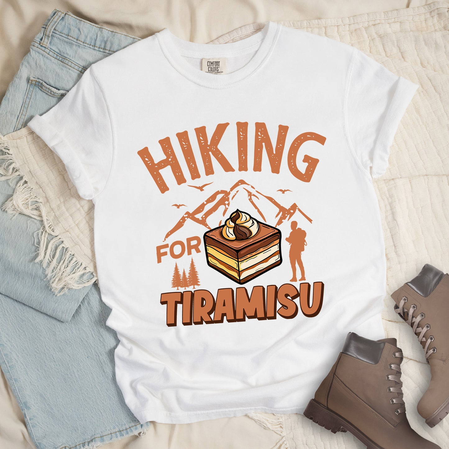 White t-shirt with light brown-colored distressed text reading "HIKING FOR TIRAMISU". The word "HIKING" appears at the top, with mountain silhouettes, small birds, and pine trees below it. A hiker silhouette stands to the right of the design. In the center is a detailed illustration of a tiramisu dessert square showing layers of coffee-soaked cake and cream, topped with a swirl of mascarpone. The word "TIRAMISU" appears in brown gradient letters at the bottom.