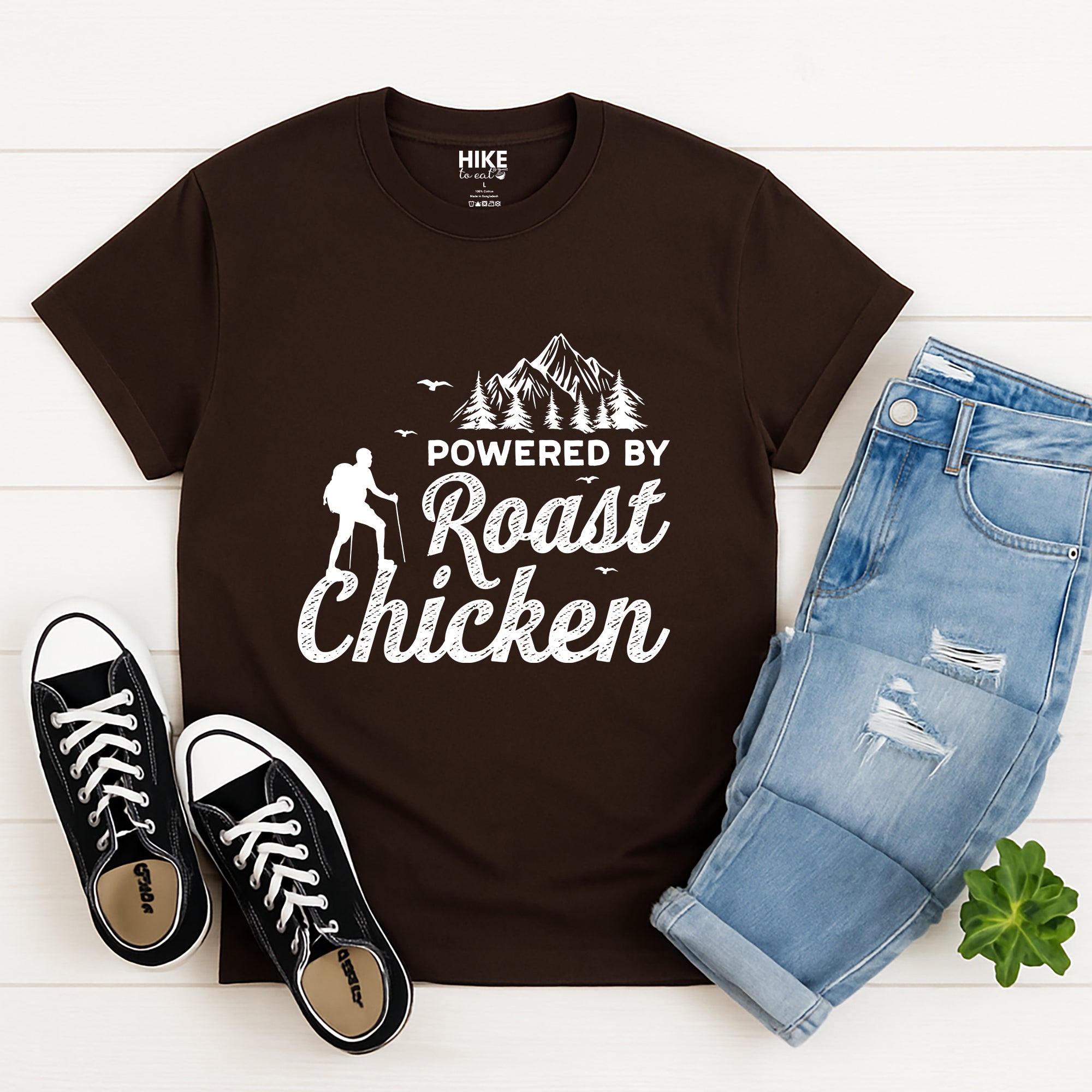 Brown t-shirt with 'Powered by Roast Chicken' design, black sneakers, and blue jeans on a white wooden surface.