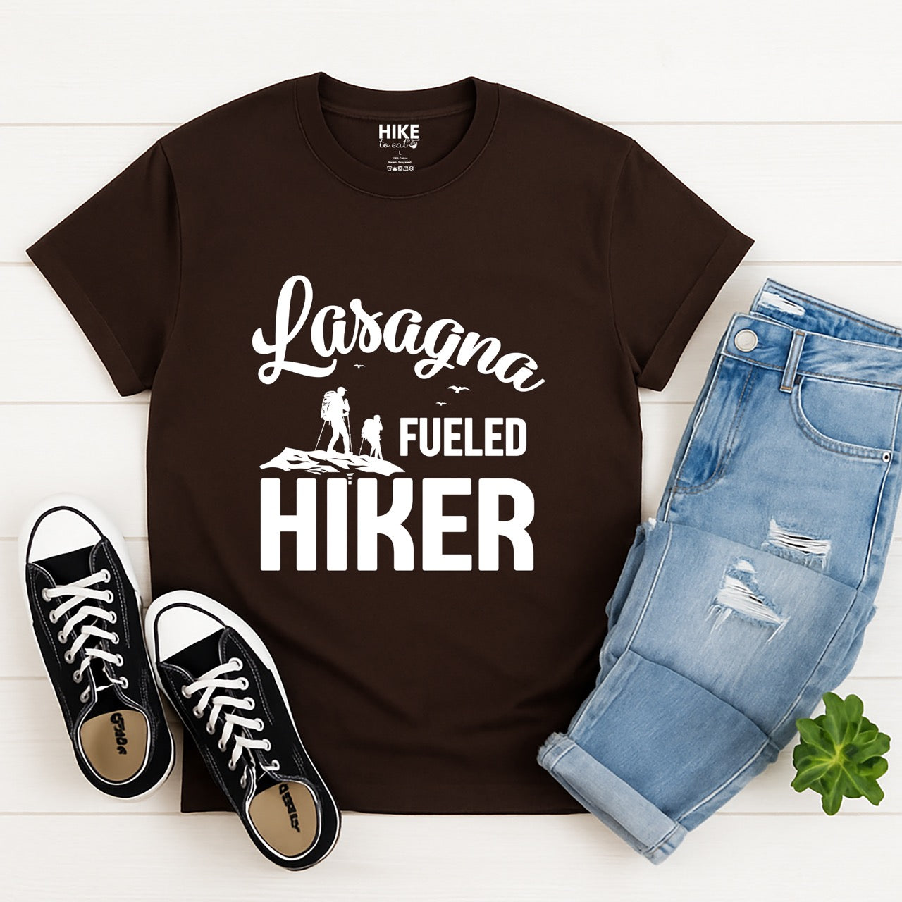 Dark Chocolate (brown) Lasagna Fueled Hiker statement tee with flowing script typography, bold lettering, two hiker silhouettes on mountain ridge, and flying birds for Italian food-loving outdoor enthusiasts, hikers and foodies laid flat with pants and shoes
