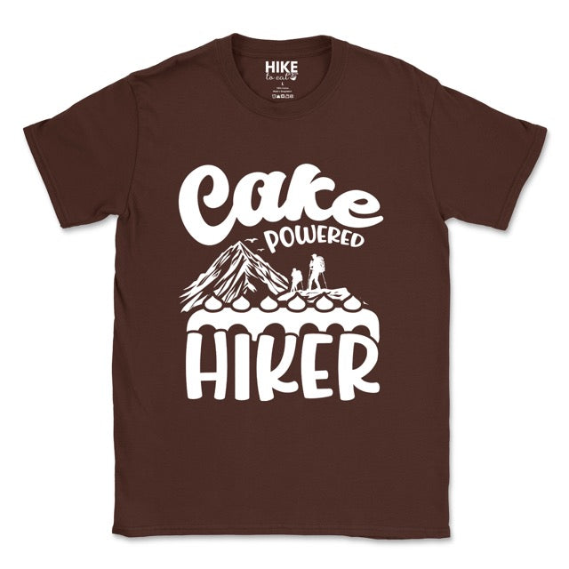 Dark chocolate Cake Powered Hiker statement tee with flowing script typography, bold lettering, mountain peaks, two hiker silhouettes, flying birds, and decorative cake illustration for dessert-loving outdoor enthusiasts, hikers and foodies