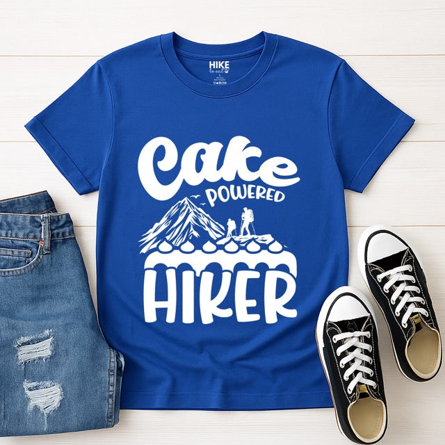 Royal Blue Cake Powered Hiker statement tee with flowing script typography, bold lettering, mountain peaks, two hiker silhouettes, flying birds, and decorative cake illustration for dessert-loving outdoor enthusiasts, hikers and foodies with shoes and pants