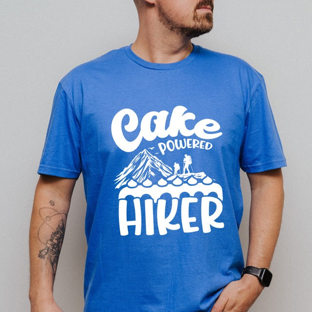 Man wearing Royal Blue Cake Powered Hiker statement tee with flowing script typography, bold lettering, mountain peaks, two hiker silhouettes, flying birds, and decorative cake illustration for dessert-loving outdoor enthusiasts, hikers and foodies