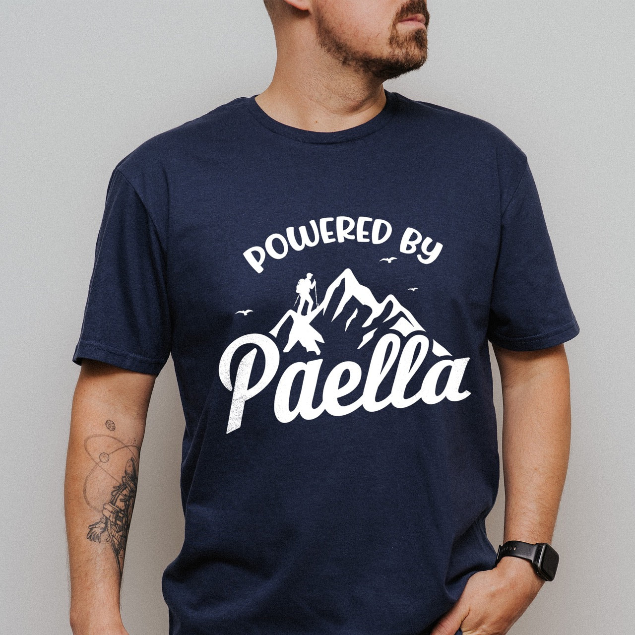 Man wearing Navy Powered by Paella statement tee with bold curved typography, flowing script lettering, mountain peaks, hiker silhouette, and flying birds for Spanish food-loving outdoor enthusiasts, hikers and foodies