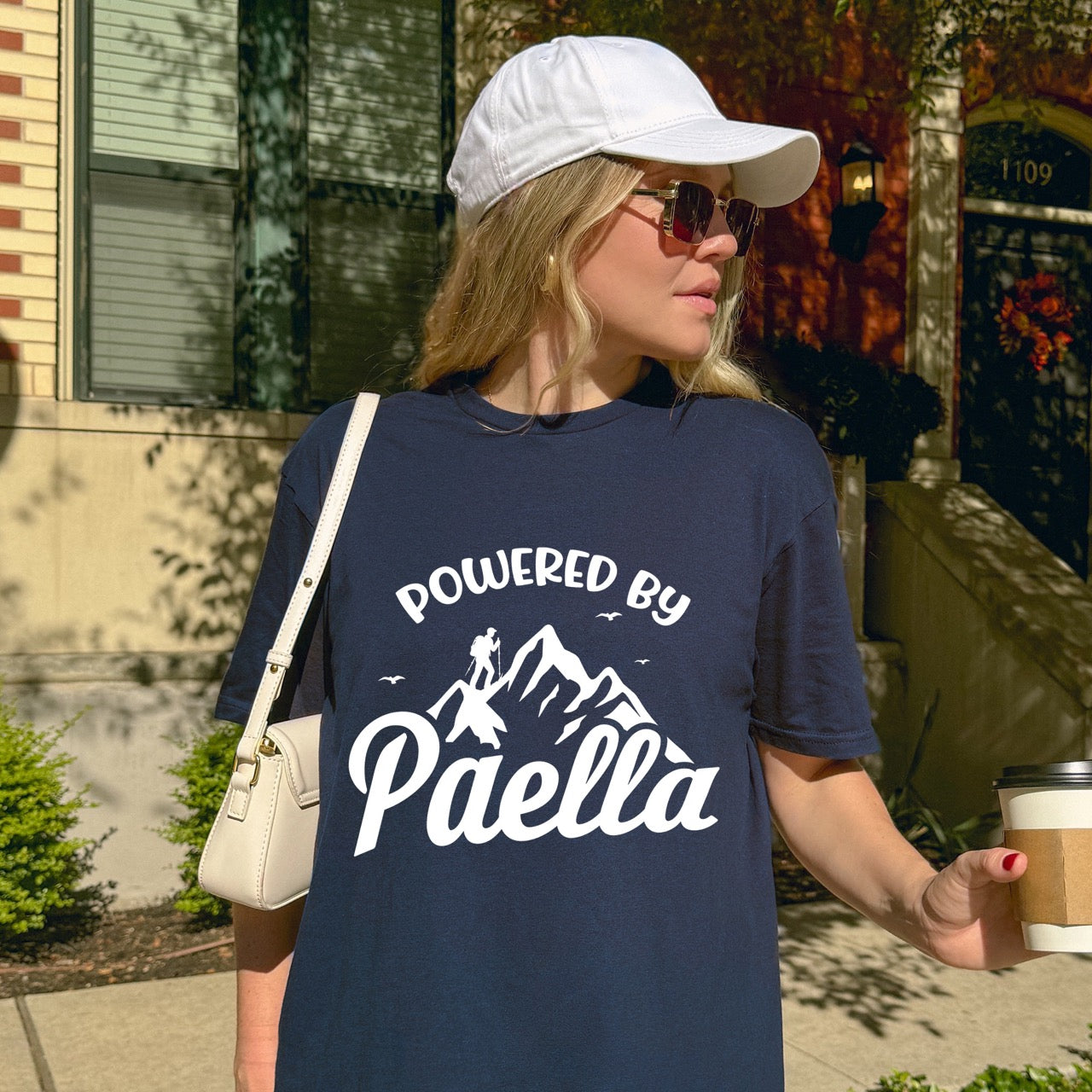 Woman wearing Navy Powered by Paella statement tee with bold curved typography, flowing script lettering, mountain peaks, hiker silhouette, and flying birds for Spanish food-loving outdoor enthusiasts, hikers and foodies