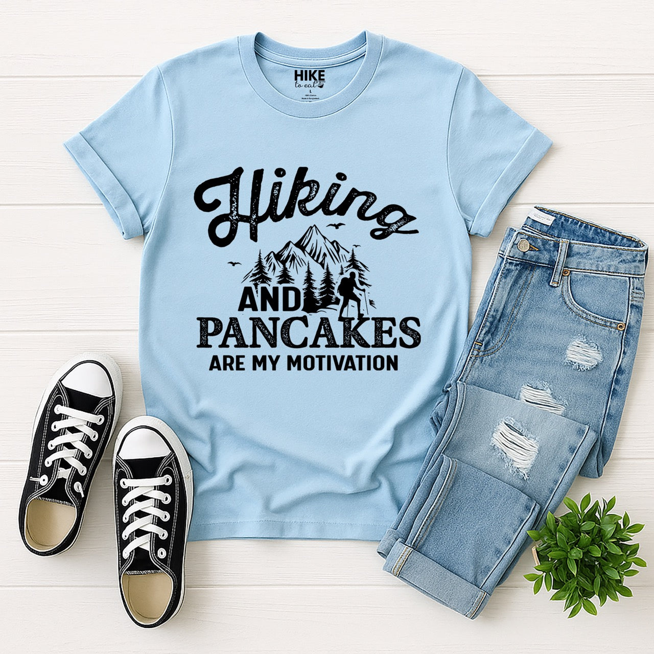 Light Blue Hiking and Pancakes are my motivation statement tee with flowing script typography, bold lettering, mountain peaks, pine trees, hiker silhouette, and flying birds for pancake-loving outdoor enthusiasts, hikers and foodies laid flat with jeans and shoes
