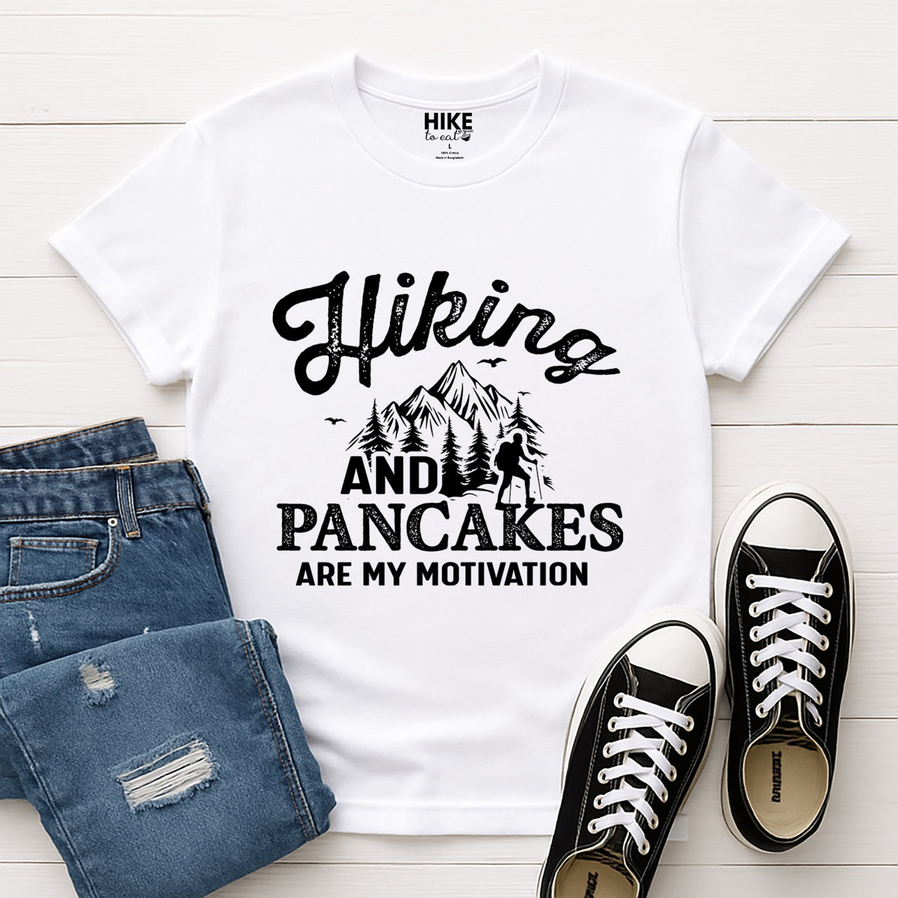 white Blue Hiking and Pancakes are my motivation statement tee with flowing script typography, bold lettering, mountain peaks, pine trees, hiker silhouette, and flying birds for pancake-loving outdoor enthusiasts, hikers and foodies laid flat with shoes and pants