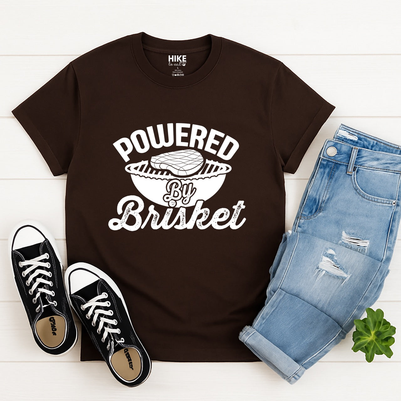 Chocolate Brown Powered by Brisket statement tee with bold curved typography, flowing script lettering, and BBQ grill with brisket illustration for barbecue-loving outdoor enthusiasts, hikers and foodies laid out with pants and shoes