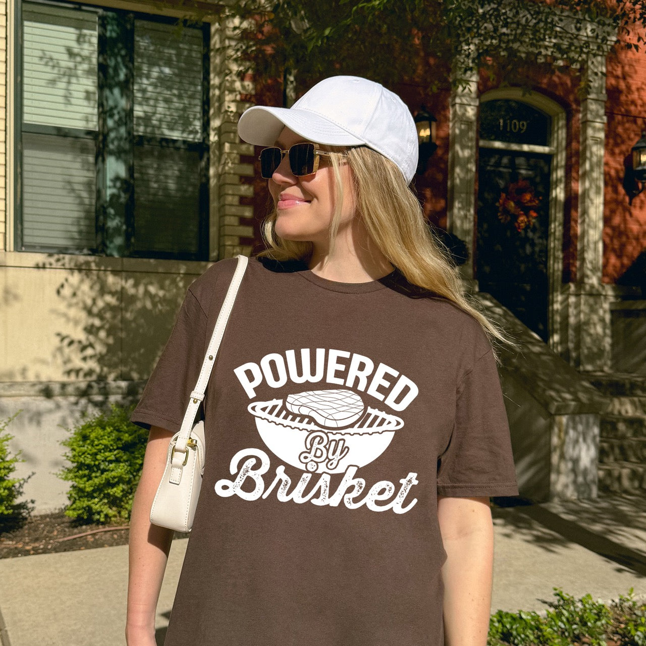 Woman wearing Chocolate Brown Powered by Brisket statement tee with bold curved typography, flowing script lettering, and BBQ grill with brisket illustration for barbecue-loving outdoor enthusiasts, hikers and foodies