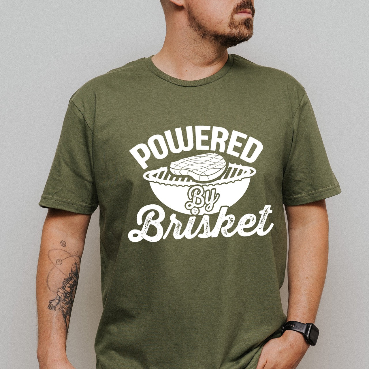 Man wearing Military Green Powered by Brisket statement tee with bold curved typography, flowing script lettering, and BBQ grill with brisket illustration for barbecue-loving outdoor enthusiasts, hikers and foodies