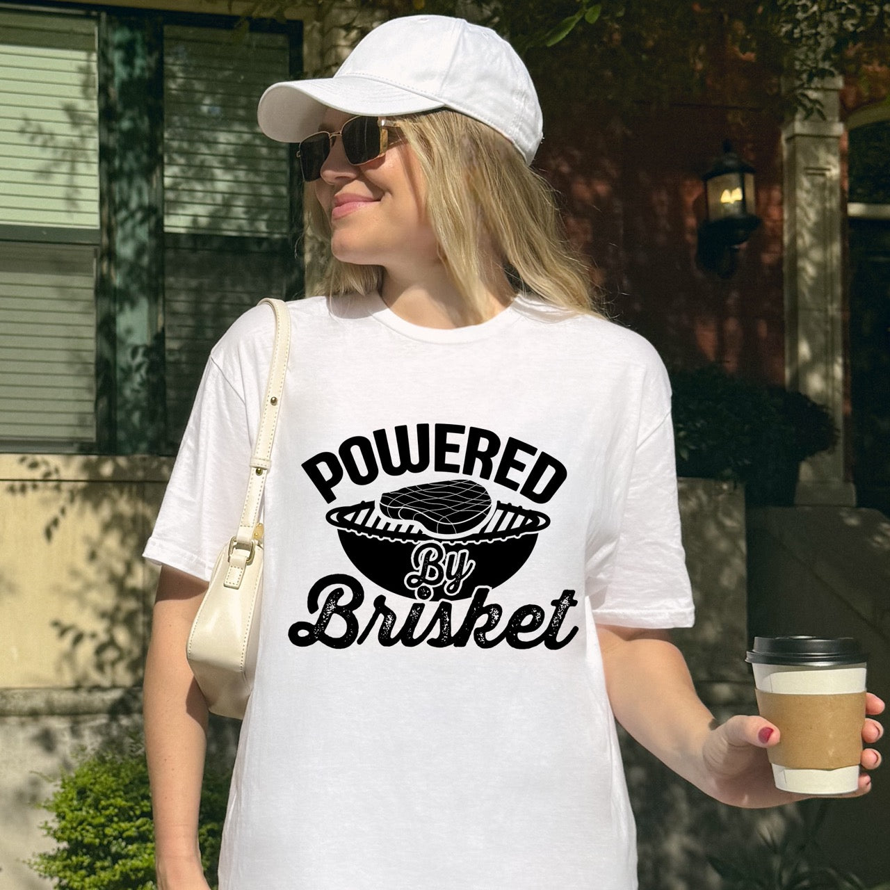 Woman wearing White  Powered by Brisket statement tee with bold curved typography, flowing script lettering, and BBQ grill with brisket illustration for barbecue-loving outdoor enthusiasts, hikers and foodies