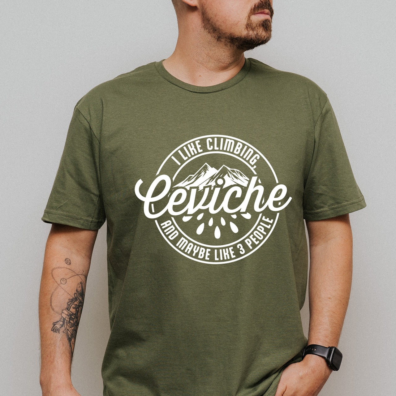 Man wearing Military Green I like Climbing, Ceviche and maybe 3 People funny introvert statement tee with circular badge design, mountain peaks, and citrus drops for seafood-loving climbers, hikers and foodies