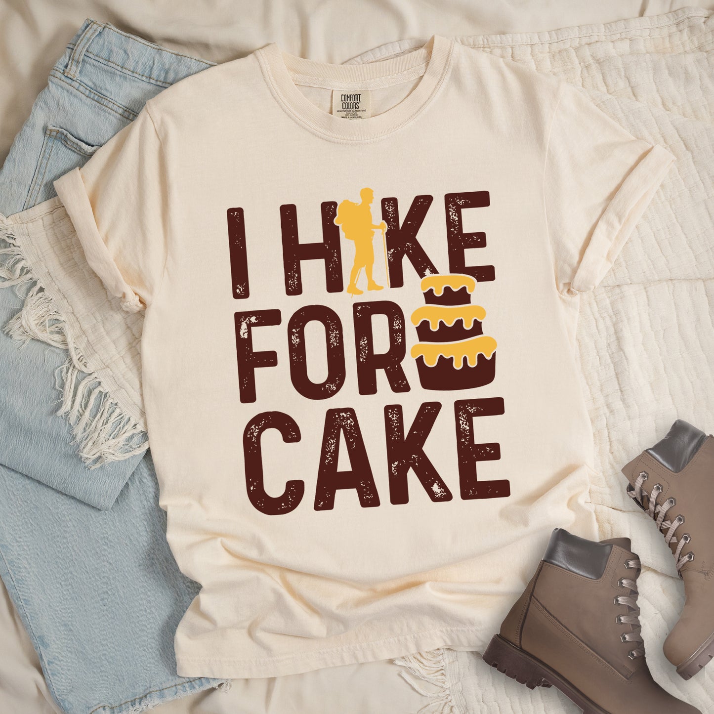 Ivory (Cream White) t-shirt with distressed deep brown-colored text reading "I HIKE FOR CAKE".  A yellow hiker silhouette is cleverly incorporated into the letter "K," while a three-tiered cake with dripping yellow icing appears between "FOR" and "CAKE." The shirt features a classic crew neck and casual rolled sleeves, styled alongside light wash denim and brown hiking boots.