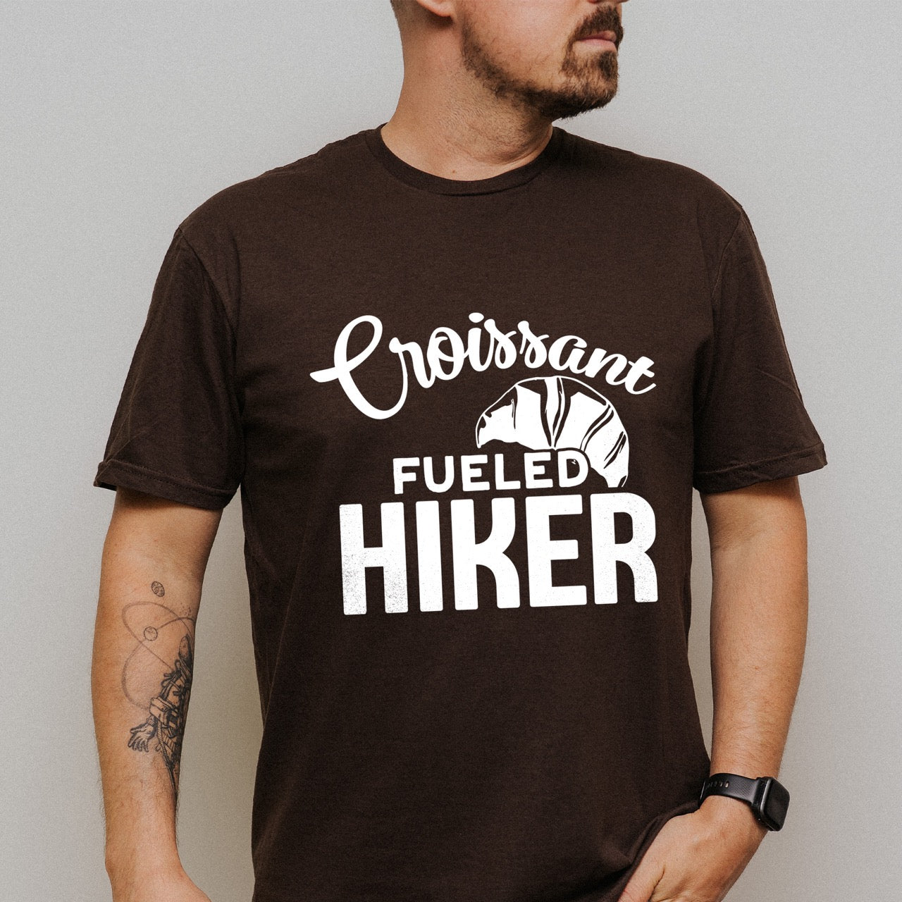 Man wearing Chocolate brown Croissant Fueled Hiker statement tee with flowing script typography, bold lettering, and croissant illustration for French pastry-loving outdoor enthusiasts, hikers and foodies