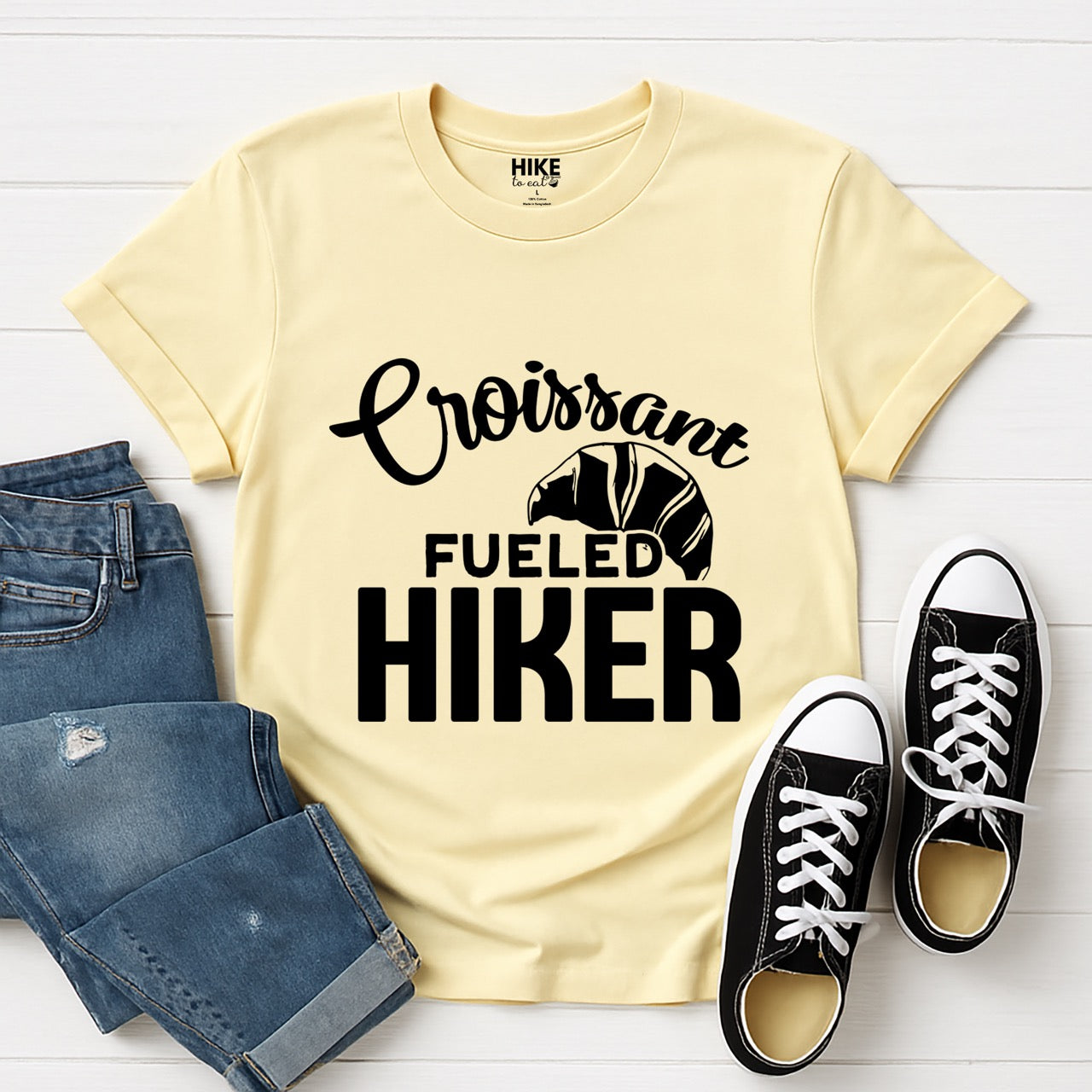Cornsilk (yellow) Croissant Fueled Hiker statement tee with flowing script typography, bold lettering, and croissant illustration for French pastry-loving outdoor enthusiasts, hikers and foodies laid flat with pants and shoes