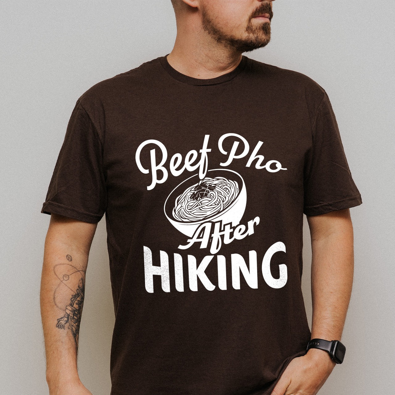 Man wearing Chocolate Brown Beef Pho After Hiking statement tee with flowing script typography, bold lettering, and detailed pho bowl illustration for Vietnamese food-loving outdoor enthusiasts, hikers and foodies