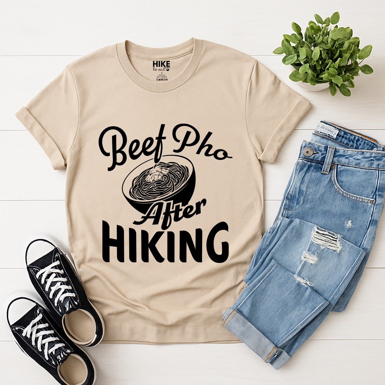 Sand (beige) Beef Pho After Hiking statement tee with flowing script typography, bold lettering, and detailed pho bowl illustration for Vietnamese food-loving outdoor enthusiasts, hikers and foodies laid flat with pants and shoes