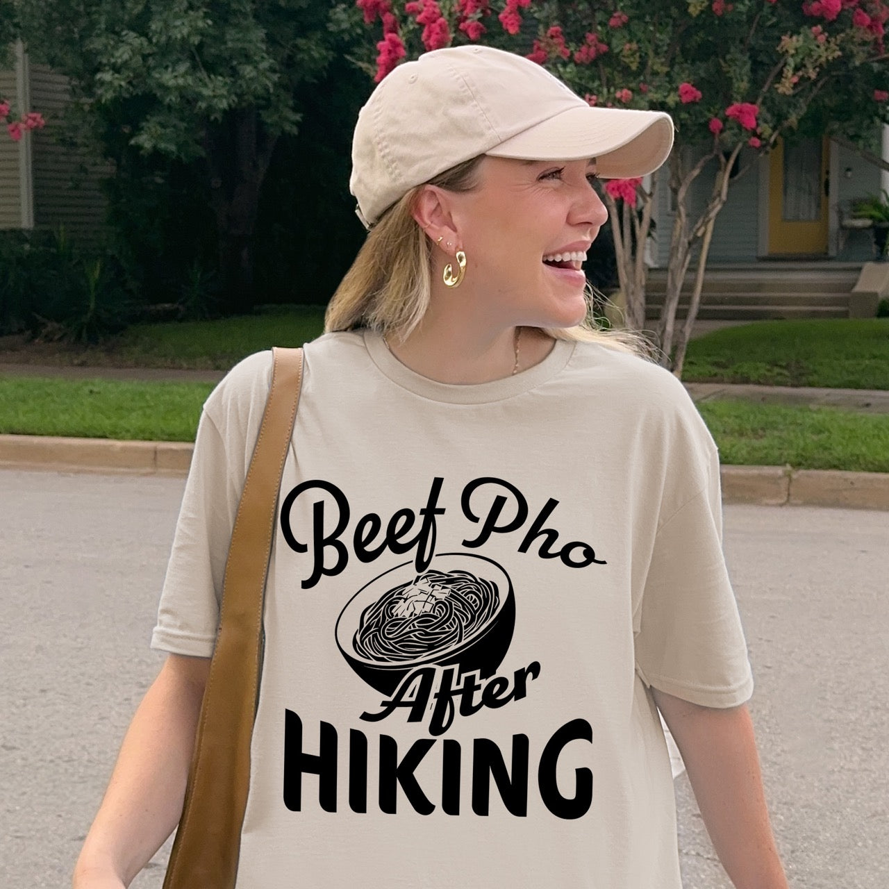Woman smiling while wearing Sand (beige) Beef Pho After Hiking statement tee with flowing script typography, bold lettering, and detailed pho bowl illustration for Vietnamese food-loving outdoor enthusiasts, hikers and foodies