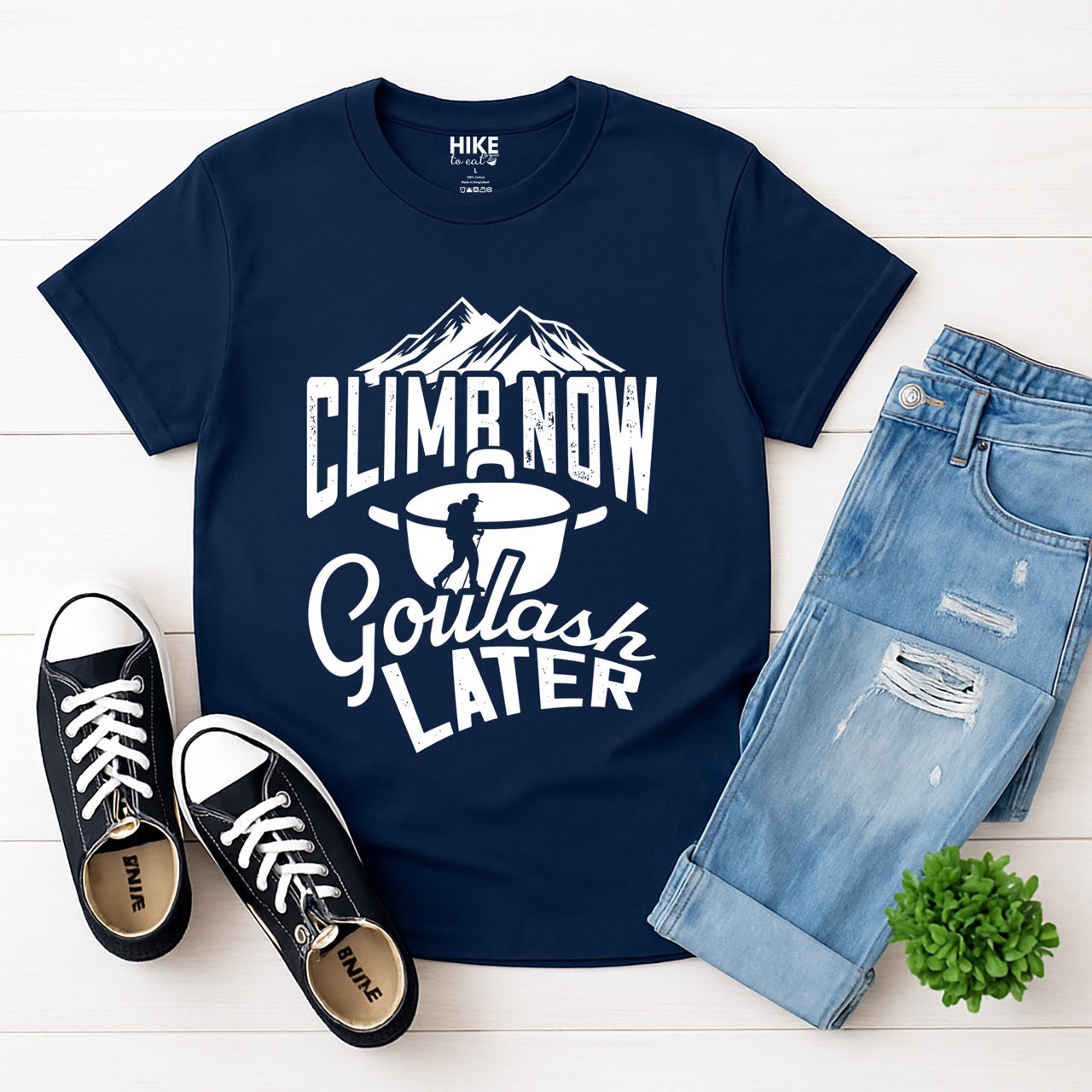 Navy Climb now Goulash Later statement tee with bold distressed typography, mountain peaks, climber silhouette, and goulash pot illustration for Hungarian food-loving climbers, hikers and foodies laid flat with pants and shoes