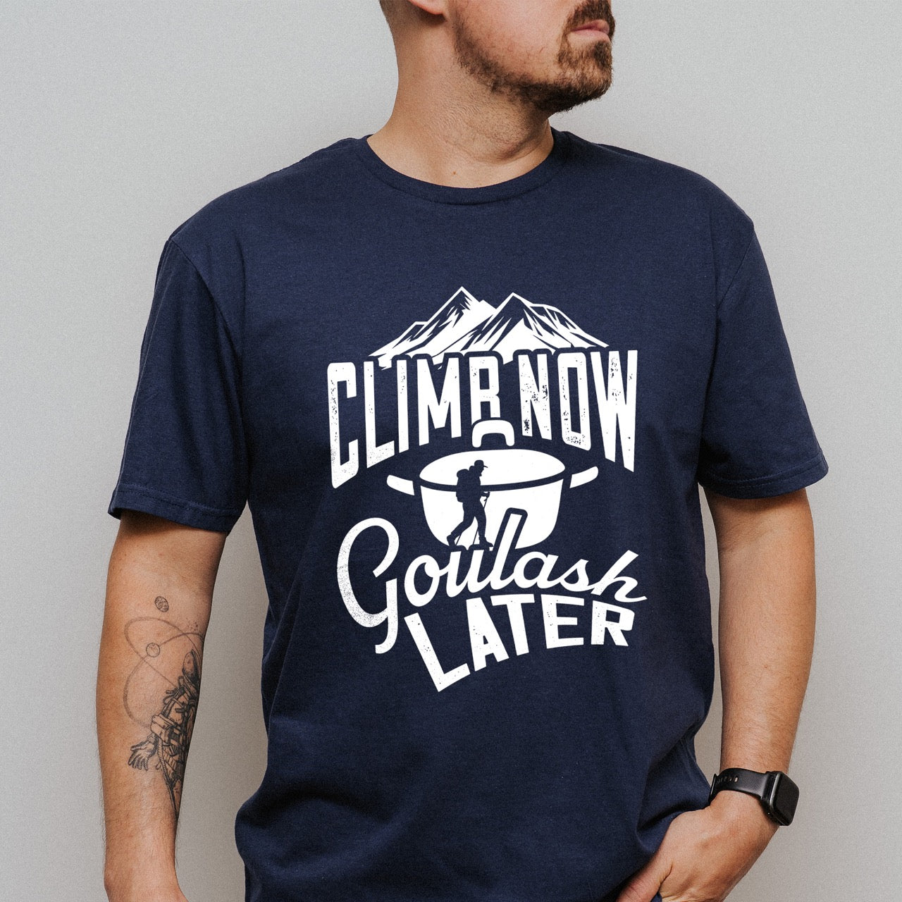 Man wearing Navy Climb now Goulash Later statement tee with bold distressed typography, mountain peaks, climber silhouette, and goulash pot illustration for Hungarian food-loving climbers, hikers and foodies
