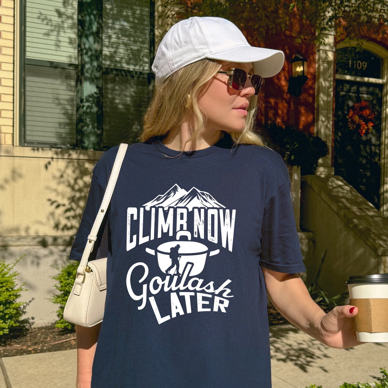 Woman wearing Navy Climb now Goulash Later statement tee with bold distressed typography, mountain peaks, climber silhouette, and goulash pot illustration for Hungarian food-loving climbers, hikers and foodies