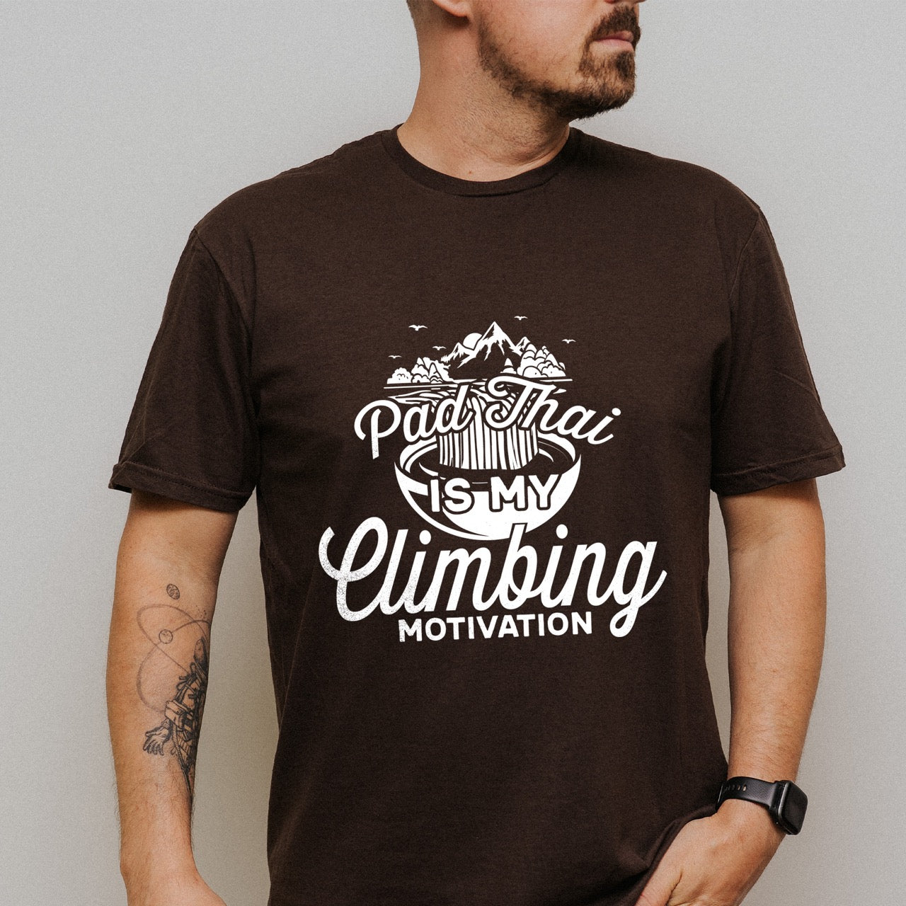 Man wearing Dark chocolate Pad Thai is my Climbing motivation statement tee with flowing script typography, mountain landscape, noodle bowl illustration, and flying birds for Thai food-loving rock climber, hikers and foodies