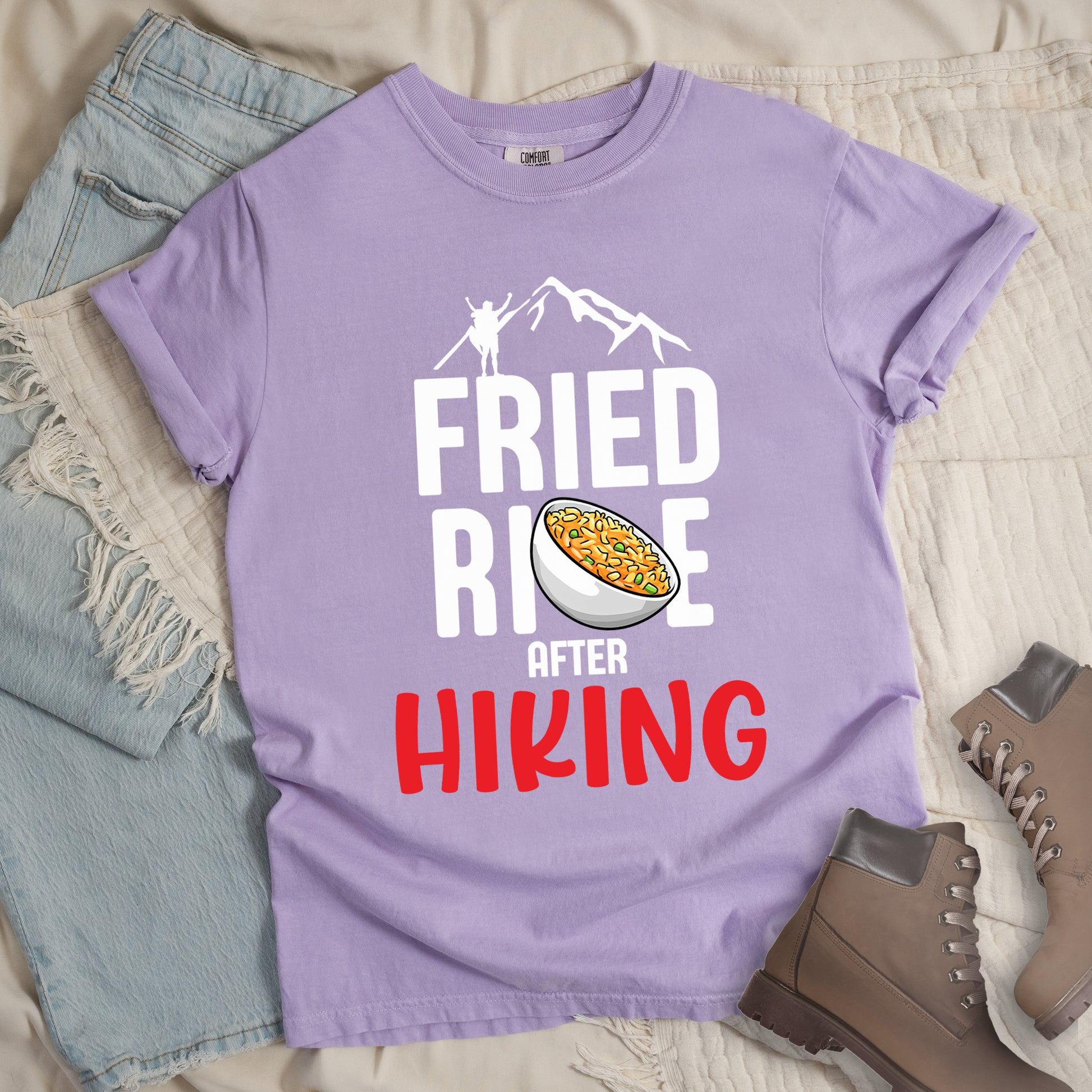 Orchid (Light Violet) t-shirt with text "FRIED RICE AFTER HIKING". The top features white mountain silhouettes with a small hiker figure. "FRIED RICE" appears in large white letters with an illustration of a bowl of fried rice replacing the letter "C" in "RICE", tilted slightly to the right. The word "AFTER" is in small white text, and "HIKING" appears at the bottom in bold red letters.