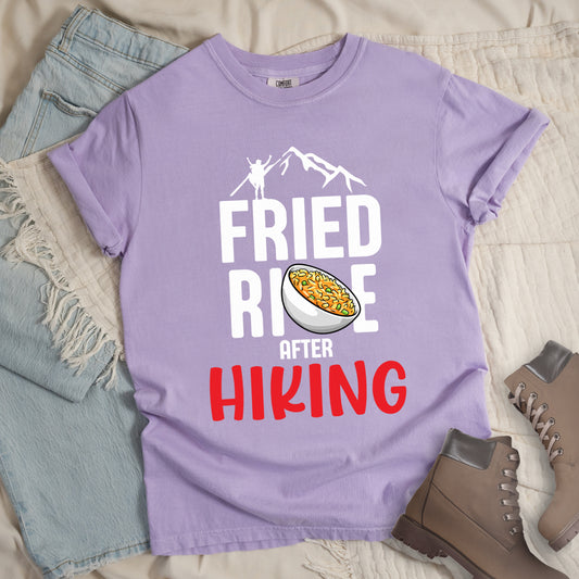 Orchid (Light Violet) t-shirt with text "FRIED RICE AFTER HIKING". The top features white mountain silhouettes with a small hiker figure. "FRIED RICE" appears in large white letters with an illustration of a bowl of fried rice replacing the letter "C" in "RICE", tilted slightly to the right. The word "AFTER" is in small white text, and "HIKING" appears at the bottom in bold red letters.