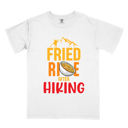 White t-shirt with text "FRIED RICE AFTER HIKING". The top features yellow mountain silhouettes with a small hiker figure. "FRIED RICE" appears in large yellow and orange letters with an illustration of a bowl of fried rice replacing the letter "C" in "RICE", tilted slightly to the right. The word "AFTER" is in small yellow text, and "HIKING" appears at the bottom in bold red letters.