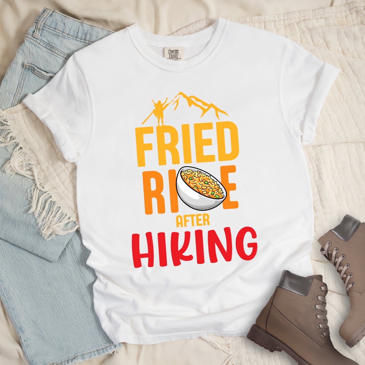 White t-shirt with text "FRIED RICE AFTER HIKING". The top features yellow mountain silhouettes with a small hiker figure. "FRIED RICE" appears in large yellow and orange letters with an illustration of a bowl of fried rice replacing the letter "C" in "RICE", tilted slightly to the right. The word "AFTER" is in small yellow text, and "HIKING" appears at the bottom in bold red letters.