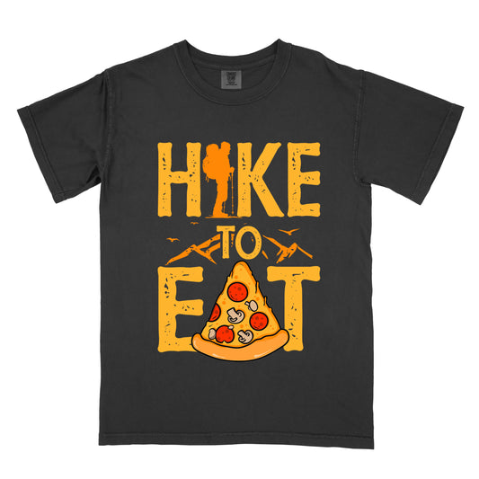 A black t-shirt featuring "HIKE TO EAT" in distressed orange-yellow lettering. The letter "I" in "HIKE" incorporates a silhouette of a hiker with a backpack. Below the words "TO" is an illustration of a pizza slice with pepperoni and mushroom toppings. Small mountain silhouettes appear under the word "TO" and also between the words "HIKE" and "EAT", with flying birds in the design.