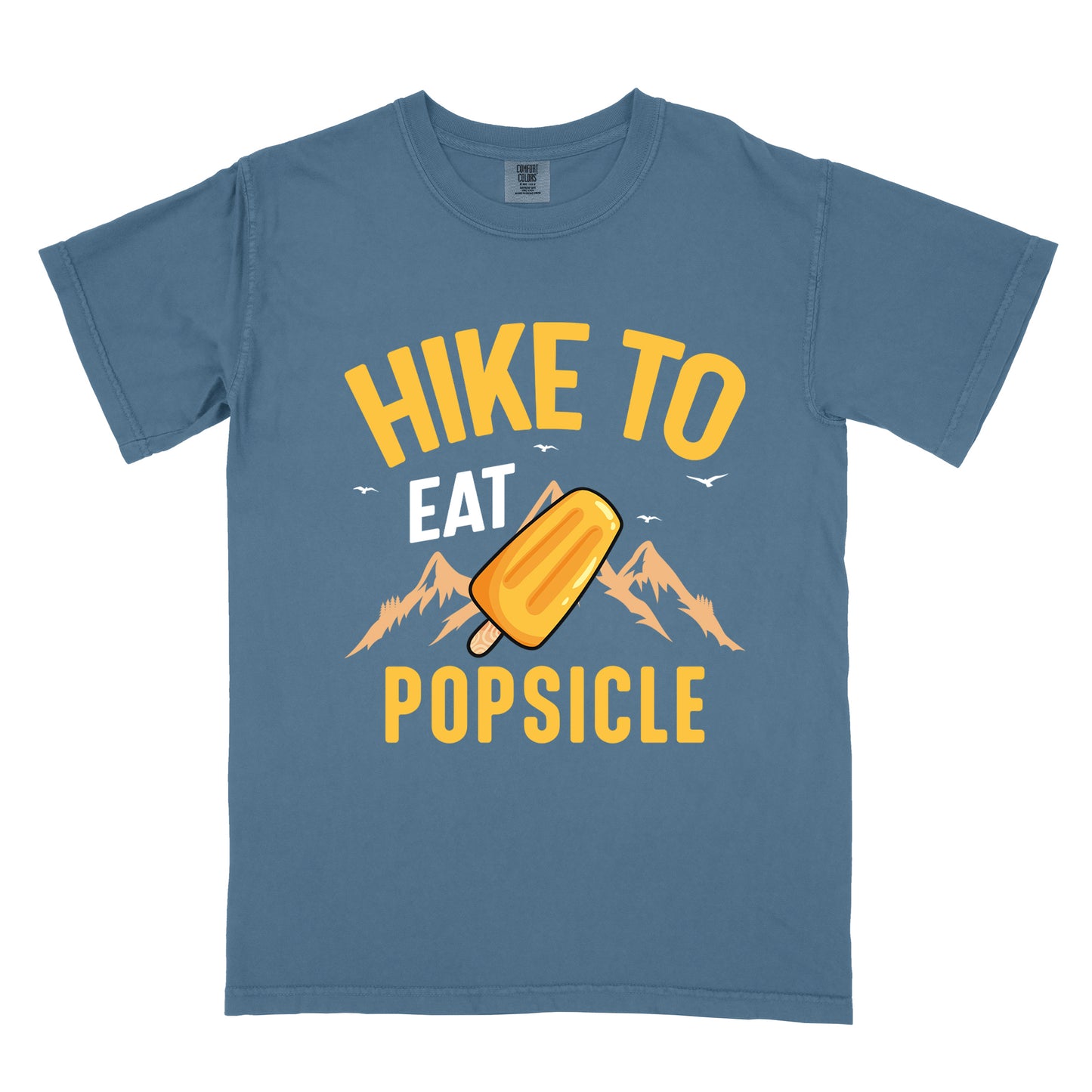 Blue Jean (Denim Blue) t-shirt with text "HIKE TO EAT POPSICLE". "HIKE TO" appears in yellow at the top, "EAT" in white in the middle, and "POPSICLE" in yellow at the bottom. Between "EAT" and "POPSICLE" is an illustration of an orange popsicle on a stick positioned in front of light orange mountain silhouettes. Small white bird silhouettes appear near the top of the design.
