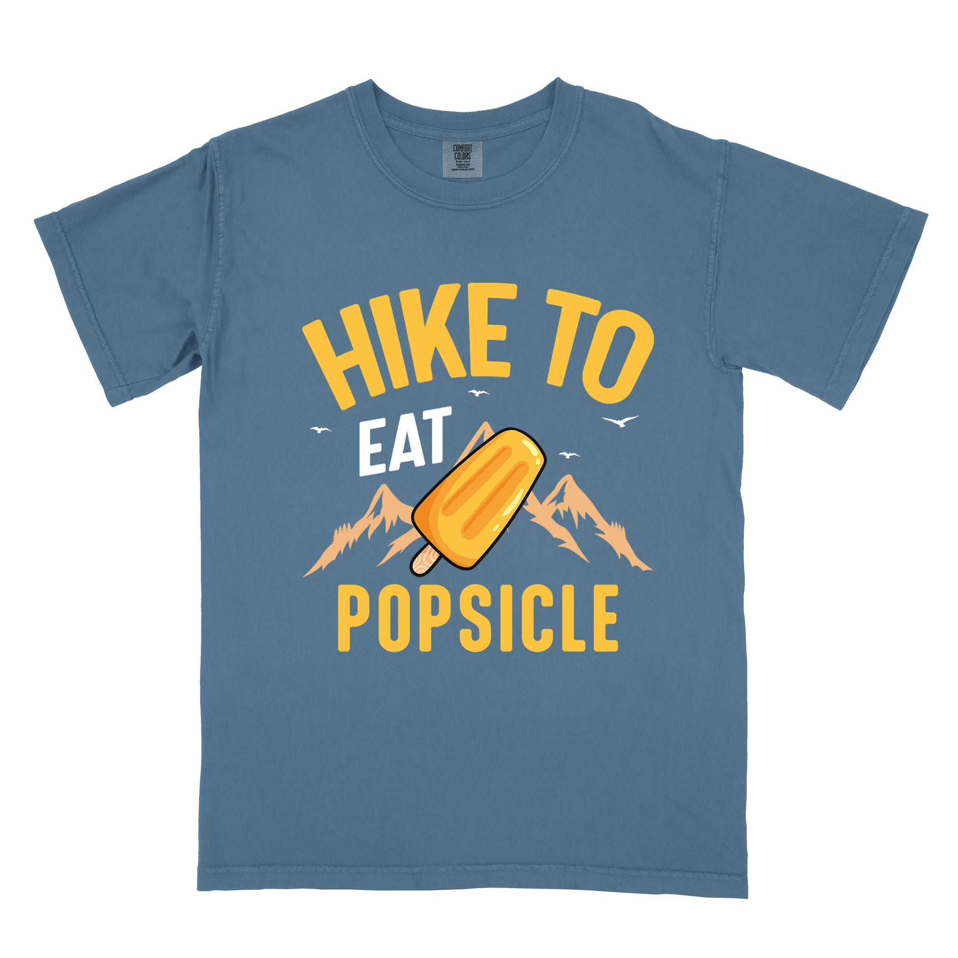 Blue Jean (Denim Blue) t-shirt with text "HIKE TO EAT POPSICLE". "HIKE TO" appears in yellow at the top, "EAT" in white in the middle, and "POPSICLE" in yellow at the bottom. Between "EAT" and "POPSICLE" is an illustration of an orange popsicle on a stick positioned in front of light orange mountain silhouettes. Small white bird silhouettes appear near the top of the design.