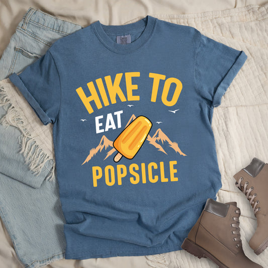 Blue Jean (Denim Blue) t-shirt with text "HIKE TO EAT POPSICLE". "HIKE TO" appears in yellow at the top, "EAT" in white in the middle, and "POPSICLE" in yellow at the bottom. Between "EAT" and "POPSICLE" is an illustration of an orange popsicle on a stick positioned in front of light orange mountain silhouettes. Small white bird silhouettes appear near the top of the design. Features a classic crew neck and casual rolled sleeves, styled alongside light wash denim and brown hiking boots. 
