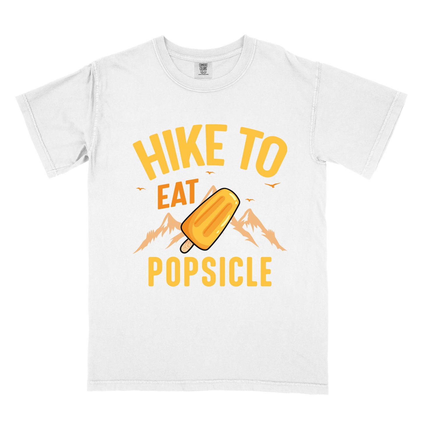 White t-shirt with text "HIKE TO EAT POPSICLE". "HIKE TO" appears in yellow at the top, "EAT" in orange in the middle, and "POPSICLE" in yellow at the bottom. Between "EAT" and "POPSICLE" is an illustration of an orange popsicle on a stick positioned in front of light orange mountain silhouettes. Small orange bird silhouettes appear near the top of the design.