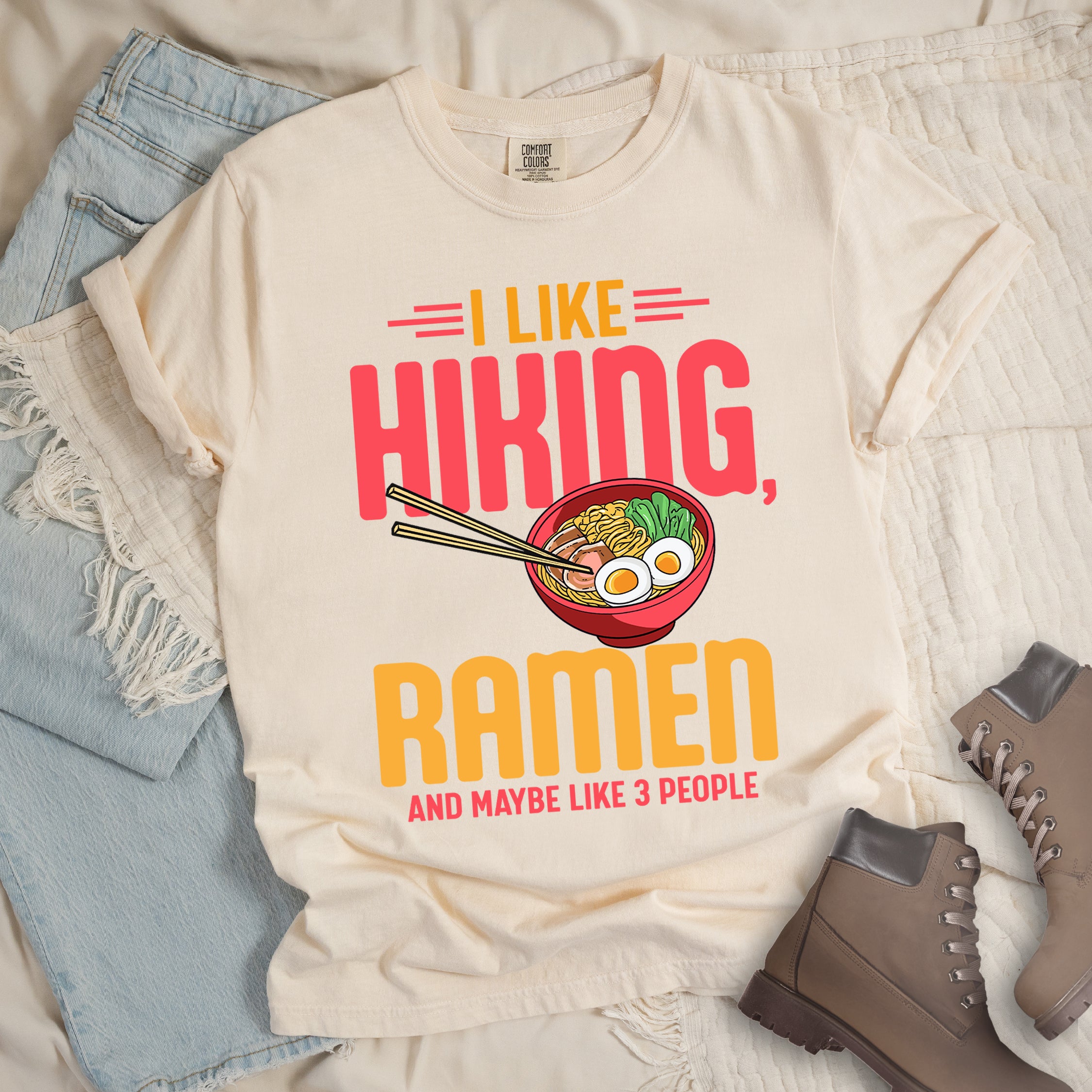 Ivory (Cream White) t-shirt with text "I LIKE HIKING, RAMEN AND MAYBE LIKE 3 PEOPLE". "I LIKE" appears in orange at the top with red decorative lines, "HIKING," in large red letters, "RAMEN" in yellow/orange, and "AND MAYBE LIKE 3 PEOPLE" in red at the bottom. Between "HIKING" and "RAMEN" is a colorful illustration of a red ramen bowl with noodles, egg, and vegetables, with chopsticks. The shirt features a classic crew neck and casual rolled sleeves, styled alongside light wash denim and brown hiking boots.