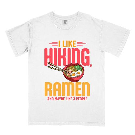 White t-shirt with text "I LIKE HIKING, RAMEN AND MAYBE LIKE 3 PEOPLE". "I LIKE" appears in orange at the top with red decorative lines, "HIKING," in large red letters, "RAMEN" in yellow/orange, and "AND MAYBE LIKE 3 PEOPLE" in red at the bottom. Between "HIKING" and "RAMEN" is a colorful illustration of a red ramen bowl with noodles, egg, and vegetables, with chopsticks.